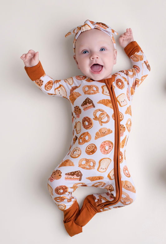 A baby smiles with arms raised, wearing the Jammie Session Doughzing Off Romper—a long-sleeve outfit with a bread and pastry print—plus a matching headband, lying on a light surface.
