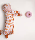 Load image into Gallery viewer, A baby wearing the Jammie Session Doughzing Off Romper and matching headband lies on a light surface, reaching for a pink frosted donut nearby.
