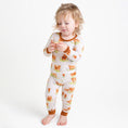 Load image into Gallery viewer, A young child in Jammie Session’s Cozy Cattle Long Sleeve PJ's—white, hypoallergenic bamboo pajamas with a brown and orange farm animal print—stands barefoot holding an object against a plain white background.