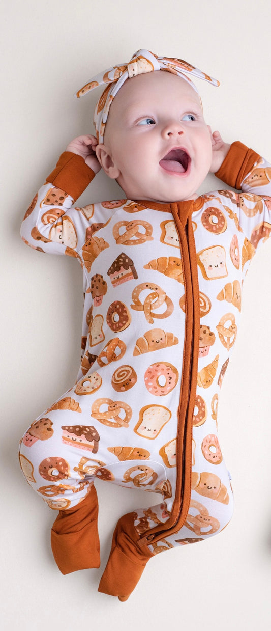 A baby smiles while wearing the Jammie Session Doughzing Off Romper, a long sleeve romper with a breakfast pastry print, and a matching headband, lying on a light surface.