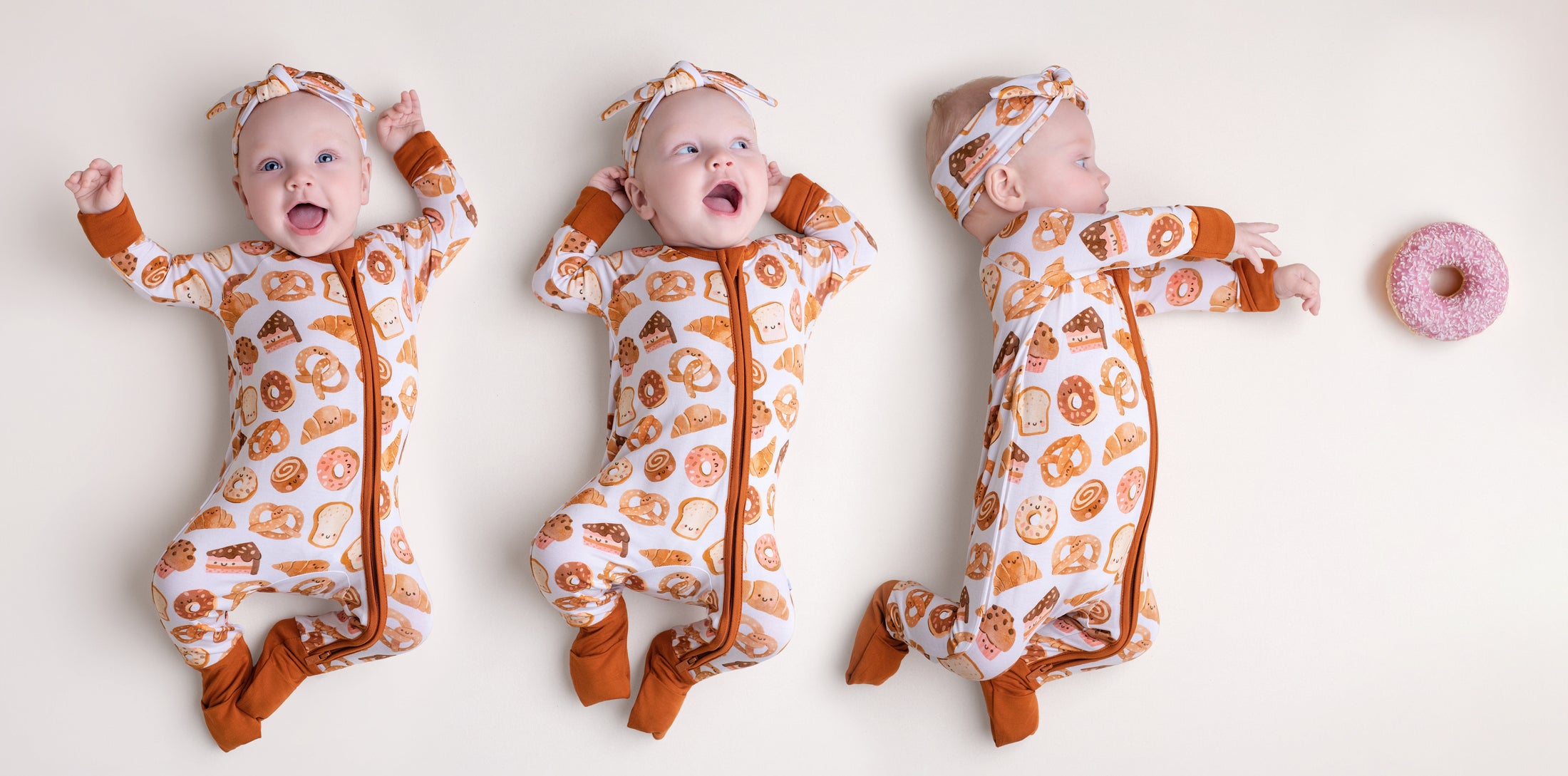 Three babies in matching Jammie Session Doughzing Off Rompers and headbands lie on a plain background, reaching for a pink frosted donut.