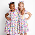 Load image into Gallery viewer, Two young girls smile side by side against a white background, wearing matching Block Party (Purple) Short Sleeve Big Kid Twirl Dresses by Big Dreams Little Jammies—soft, hypoallergenic bamboo outfits perfect for sensitive skin.