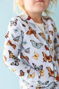 Load image into Gallery viewer, A child models the Jammie Session Butterfly Kisses Kids' Jacket—featuring colorful butterfly prints—against a light blue background.