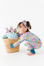 A toddler wears the Jammie Session Whispering Wildflowers Romper and headband, crouching beside a wicker basket of blue and purple faux flowers against a white background.