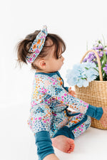 A toddler in a Jammie Session Whispering Wildflowers Romper and matching headband sits on the floor beside a wicker basket filled with artificial flowers.