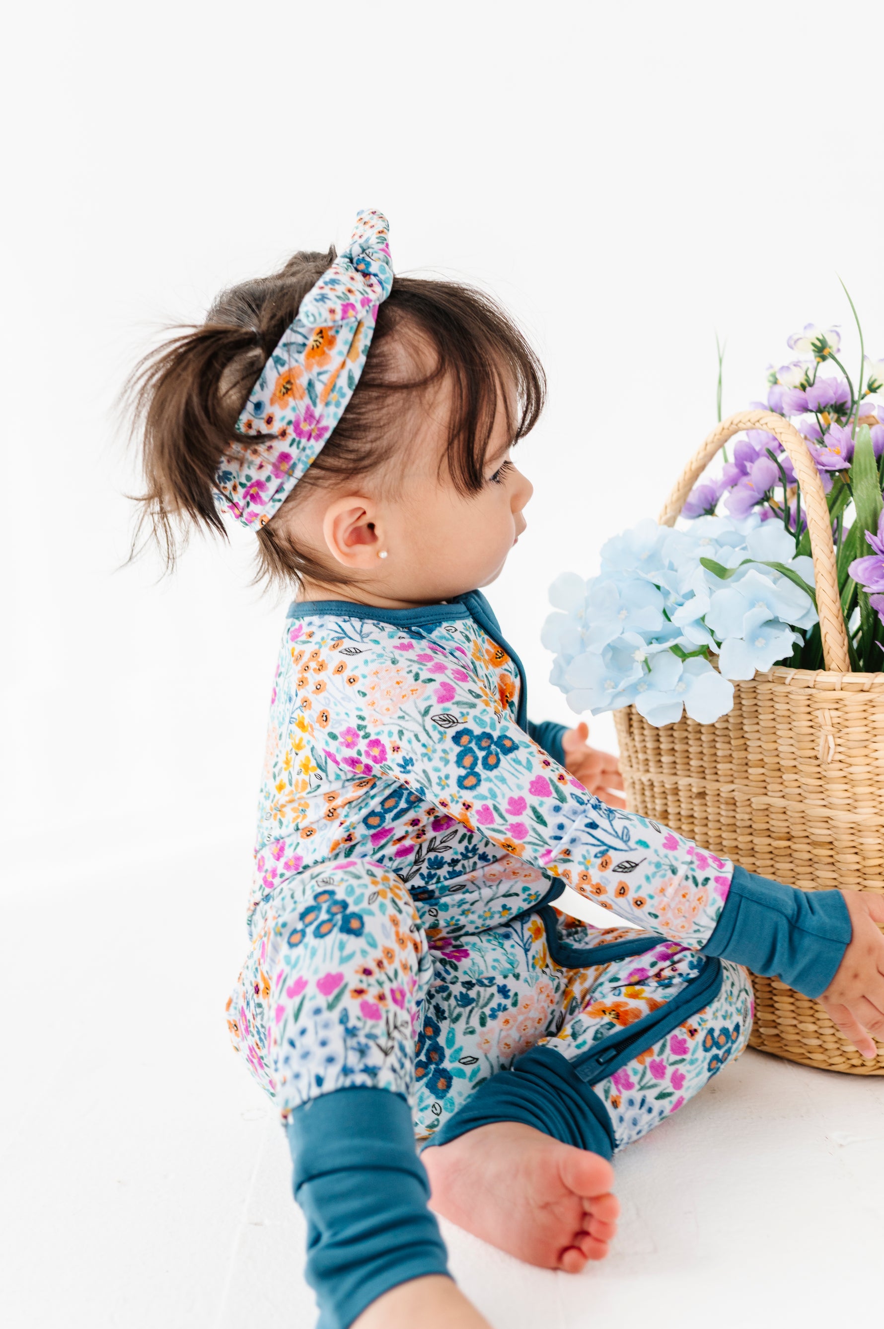 A toddler in a Jammie Session Whispering Wildflowers Romper and matching headband sits on the floor beside a wicker basket filled with artificial flowers.