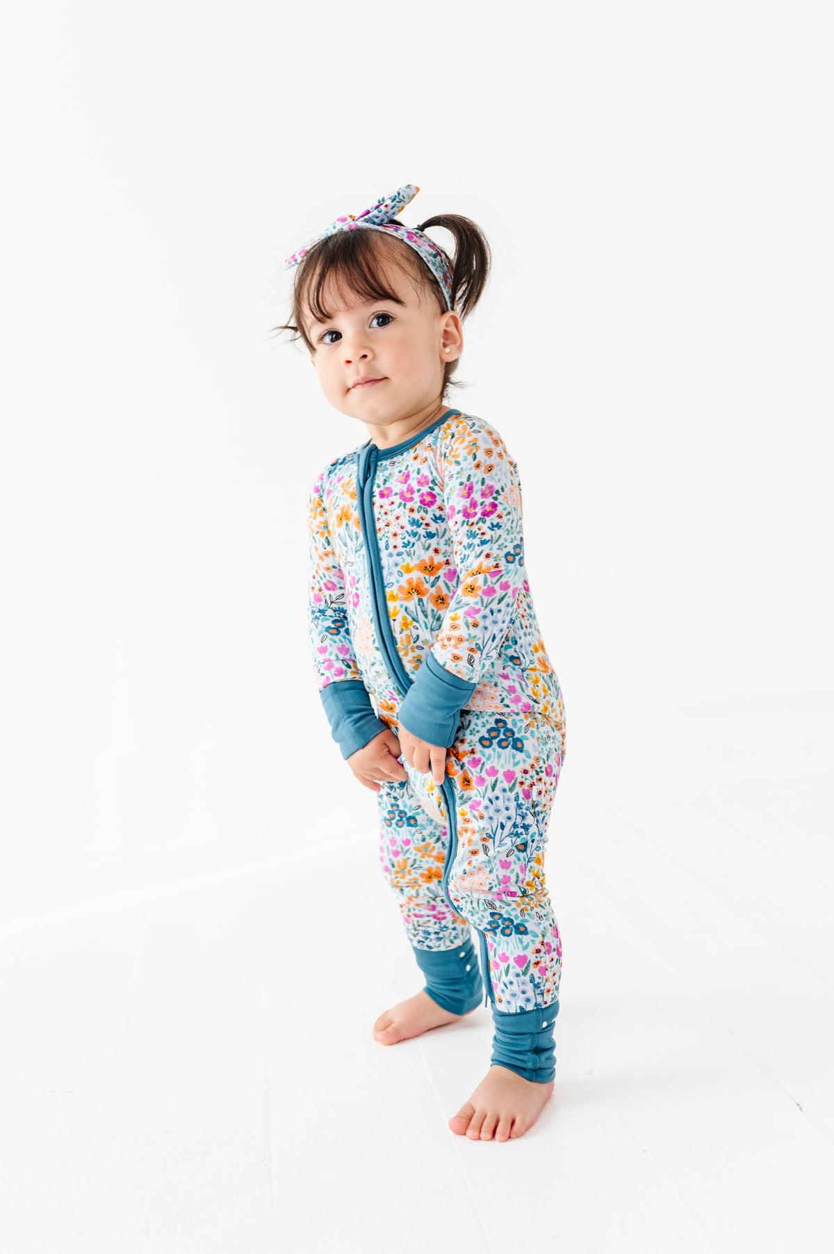 A toddler wearing the Jammie Session Whispering Wildflowers Romper with matching headband stands barefoot on a white background.