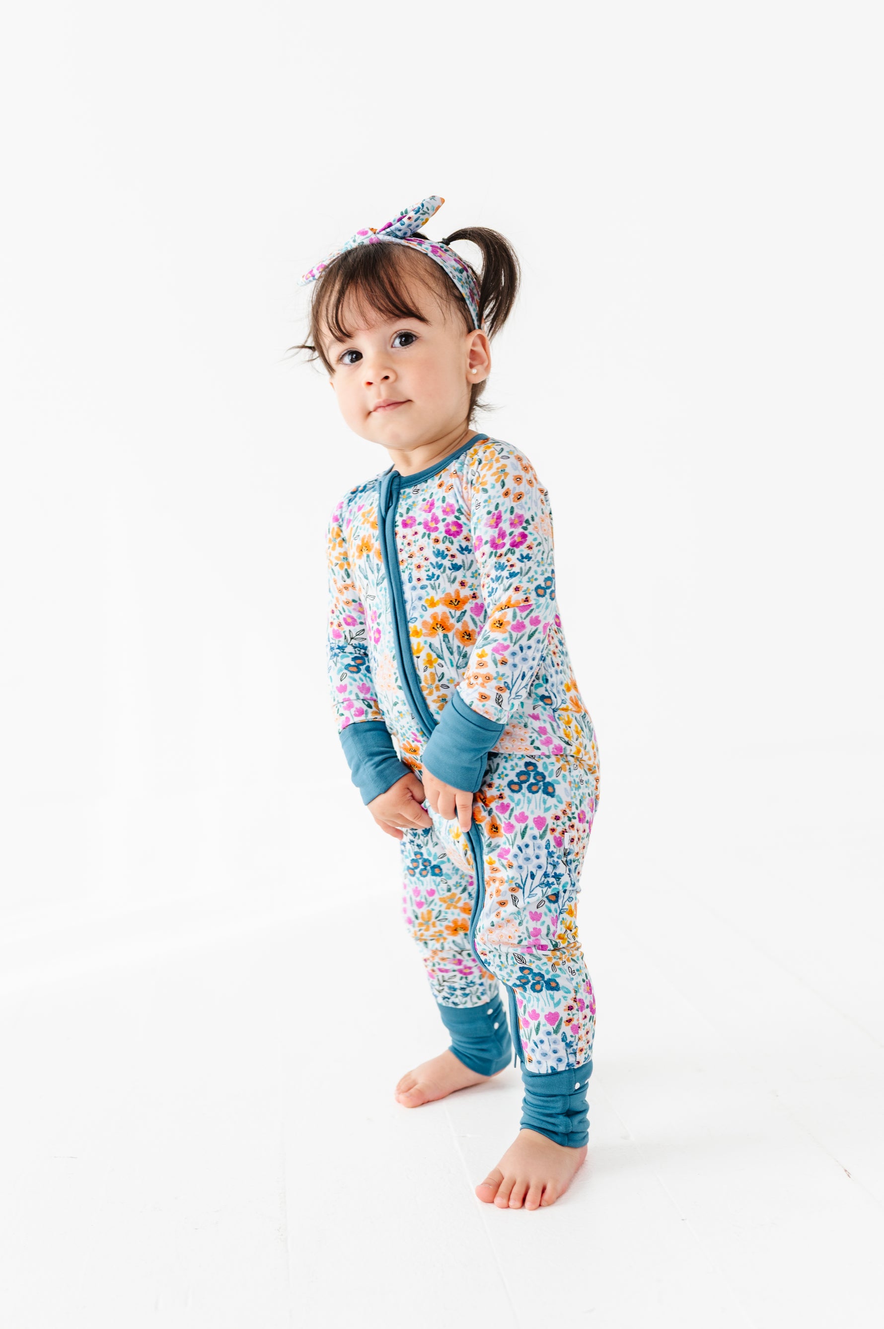 A toddler wearing the Jammie Session Whispering Wildflowers Romper with matching headband stands barefoot on a white background.