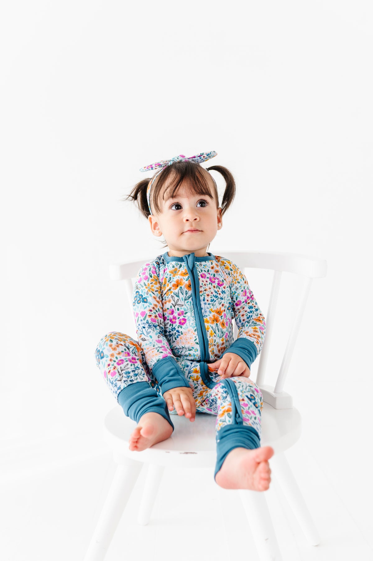 A young child with pigtails sits on a white chair, wearing the Jammie Session Whispering Wildflowers Romper and a matching bow headband.