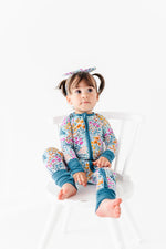 A young child with pigtails sits on a white chair, wearing the Jammie Session Whispering Wildflowers Romper and a matching bow headband.