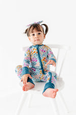 A young child with pigtails sits barefoot on a white chair, wearing the Jammie Session Whispering Wildflowers Romper and a matching headband, set against a plain white background.