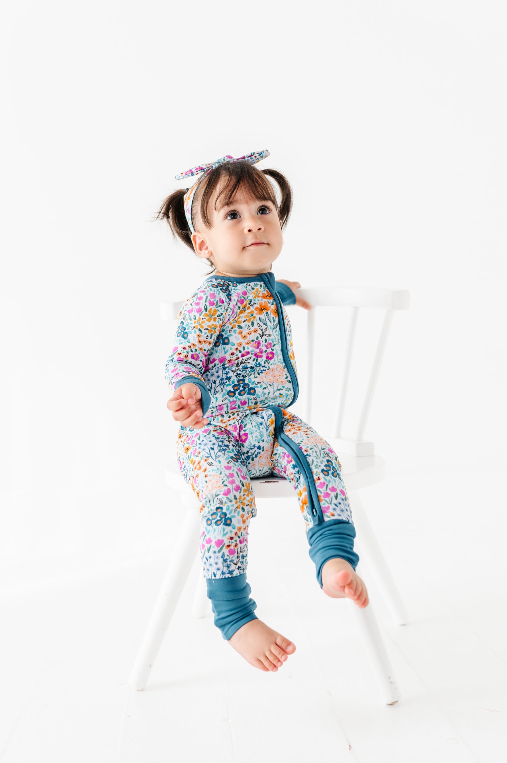 A young child with pigtails sits on a white chair, wearing the Jammie Session Whispering Wildflowers Romper and matching headband, against a plain white background.