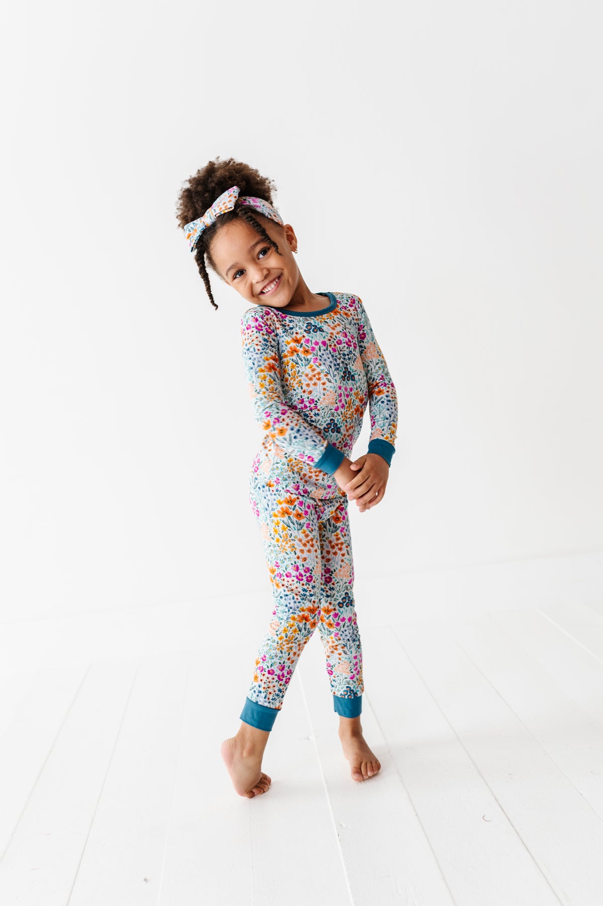 A young girl stands barefoot on a white floor, smiling in Jammie Session’s Whispering Wildflowers Long Sleeve PJ's and matching headband.