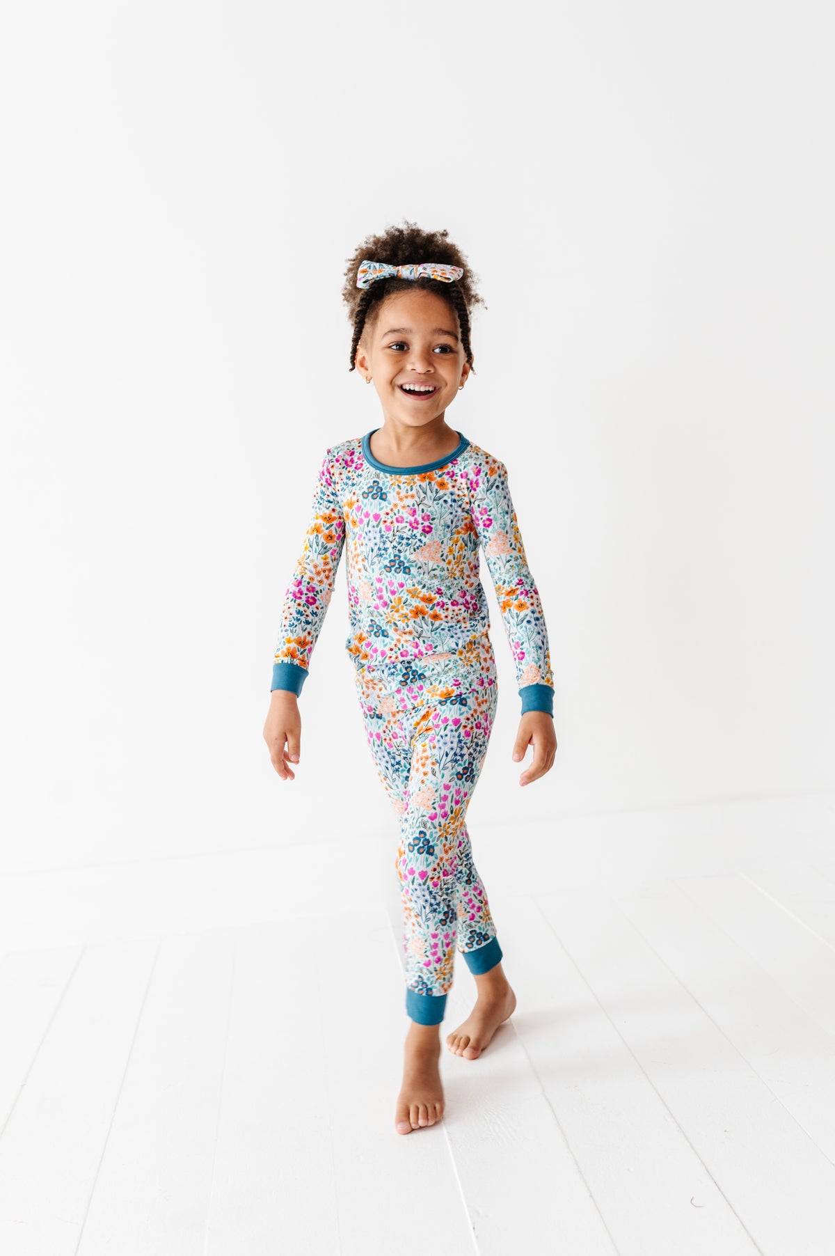 A young child models the Jammie Session Whispering Wildflowers Long Sleeve PJ's with a matching headband, walking barefoot on a white floor against a plain white background.