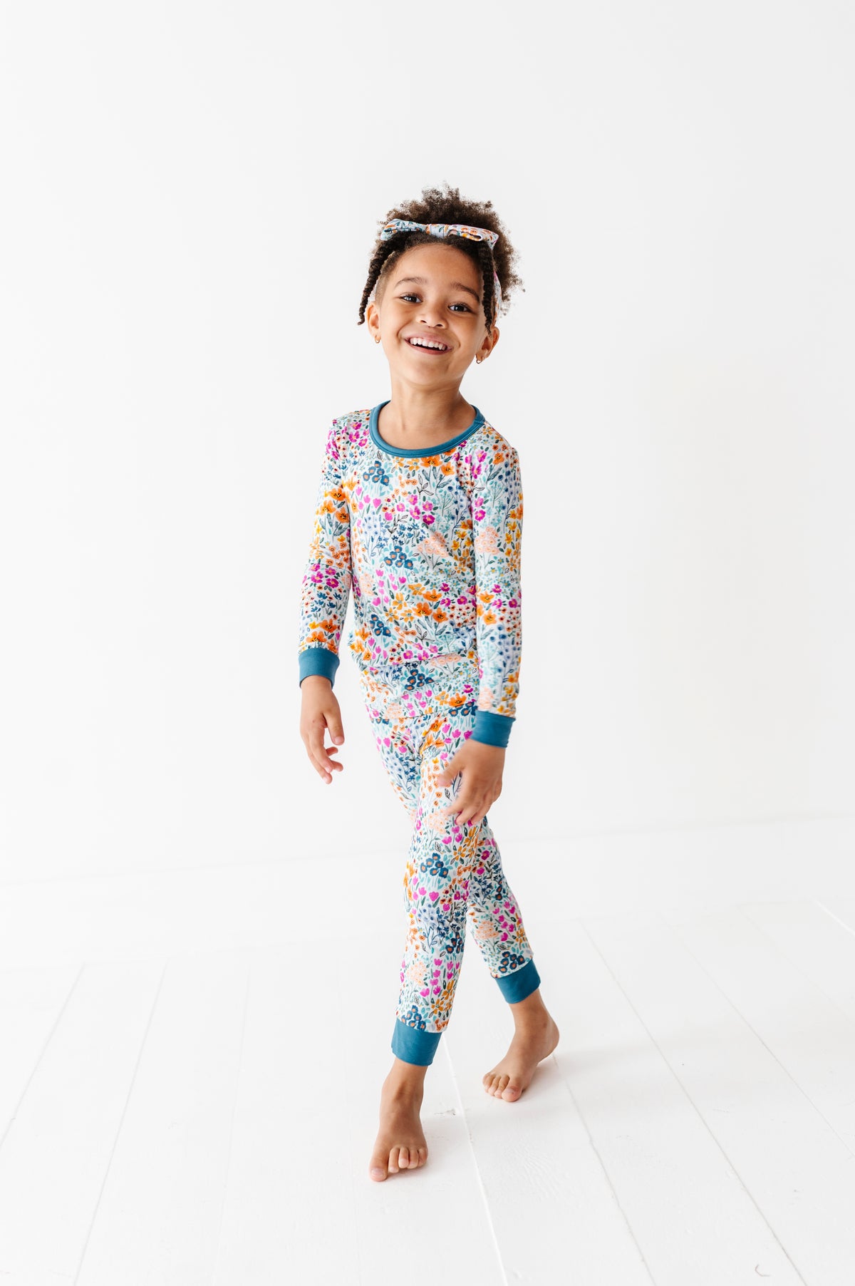 A young child smiles at the camera, barefoot on a white floor, wearing Jammie Session’s Whispering Wildflowers Long Sleeve PJ’s—colorful, floral-patterned bamboo pajamas that are hypoallergenic for comfort and style.