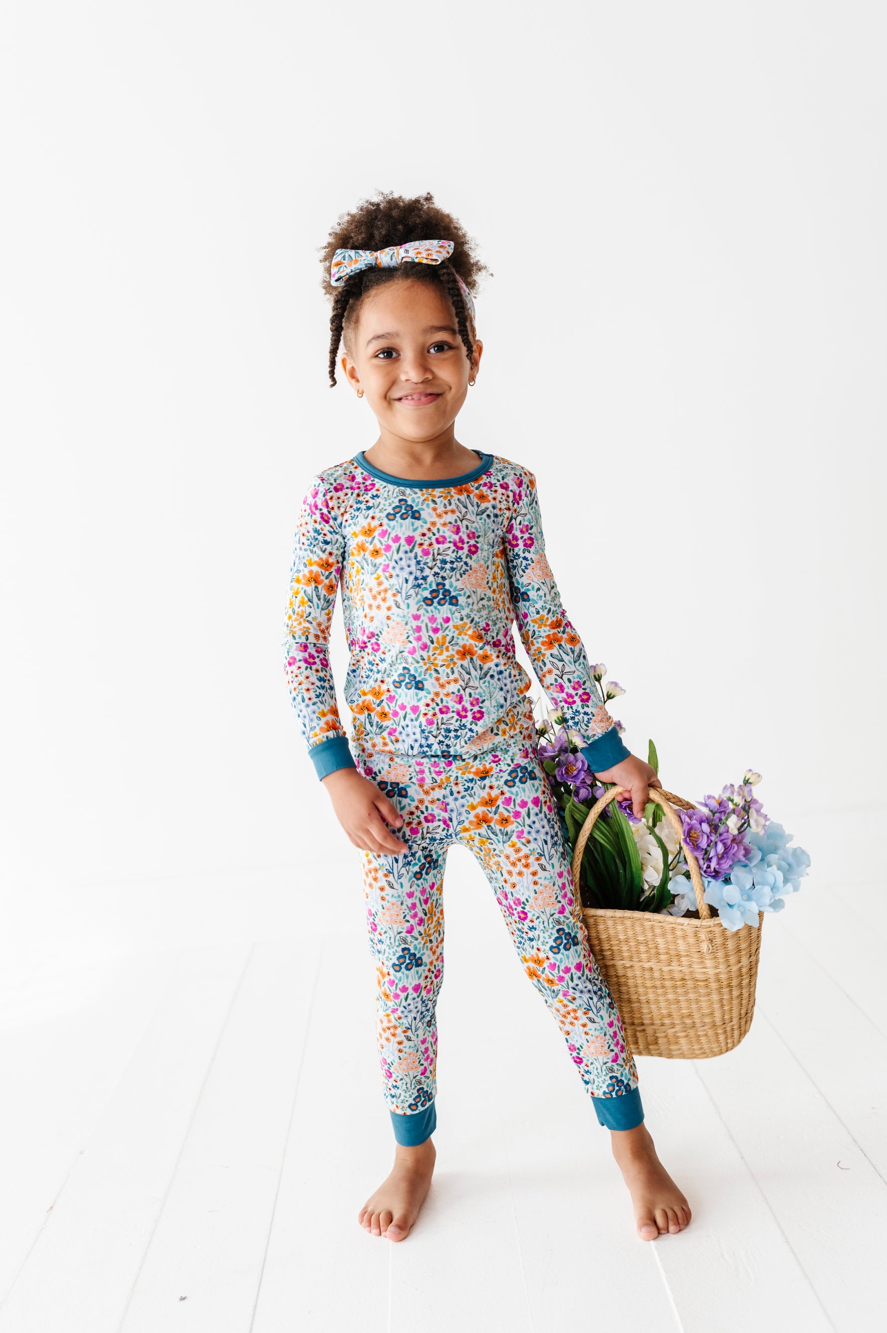 A young child stands barefoot in Jammie Session's Whispering Wildflowers Long Sleeve PJ's with a matching headband, holding a basket of colorful flowers against a white background.