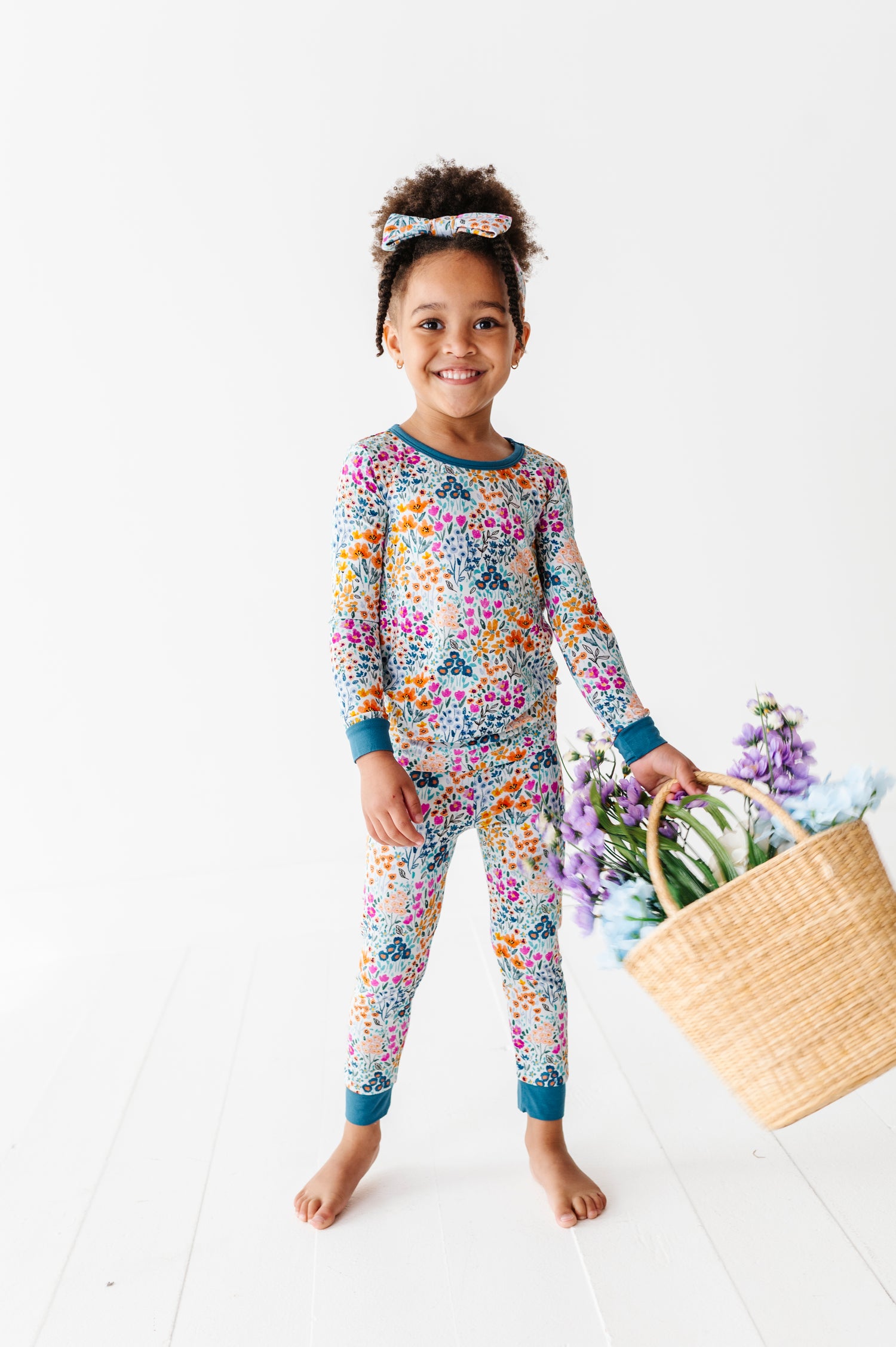 A young child wears Jammie Session's Whispering Wildflowers Long Sleeve PJ’s with a matching headband, holding a wicker basket of purple flowers, standing on a white floor against a plain white background.