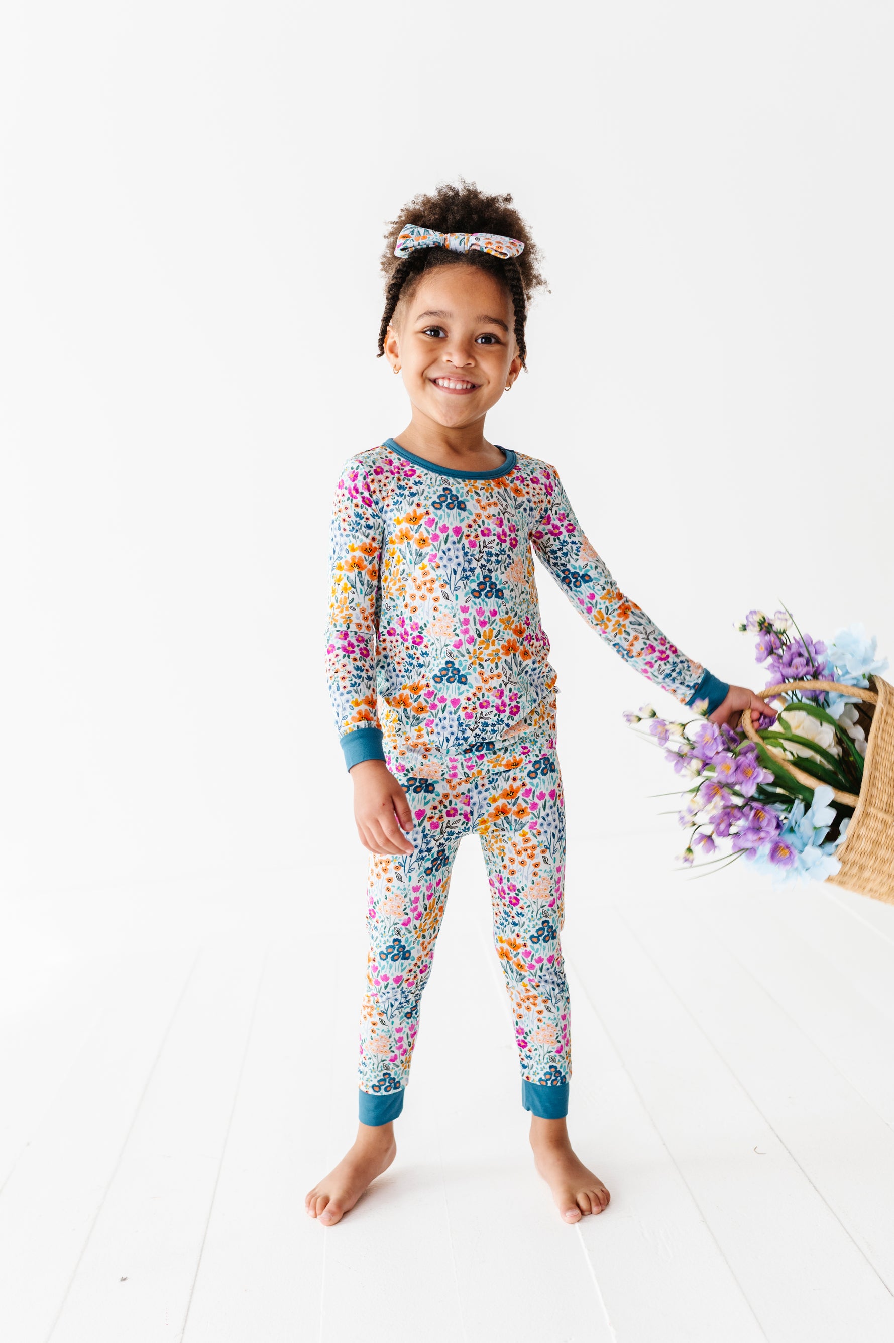 A young girl smiles barefoot on a white floor in Jammie Session’s Whispering Wildflowers Long Sleeve PJ's with a matching headband, as a basket of purple and blue flowers is tipped over nearby.