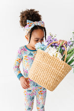 A young girl wears Jammie Session's Whispering Wildflowers Long Sleeve PJ's with a matching headband, holding a wicker basket of assorted flowers, standing against a plain white background.