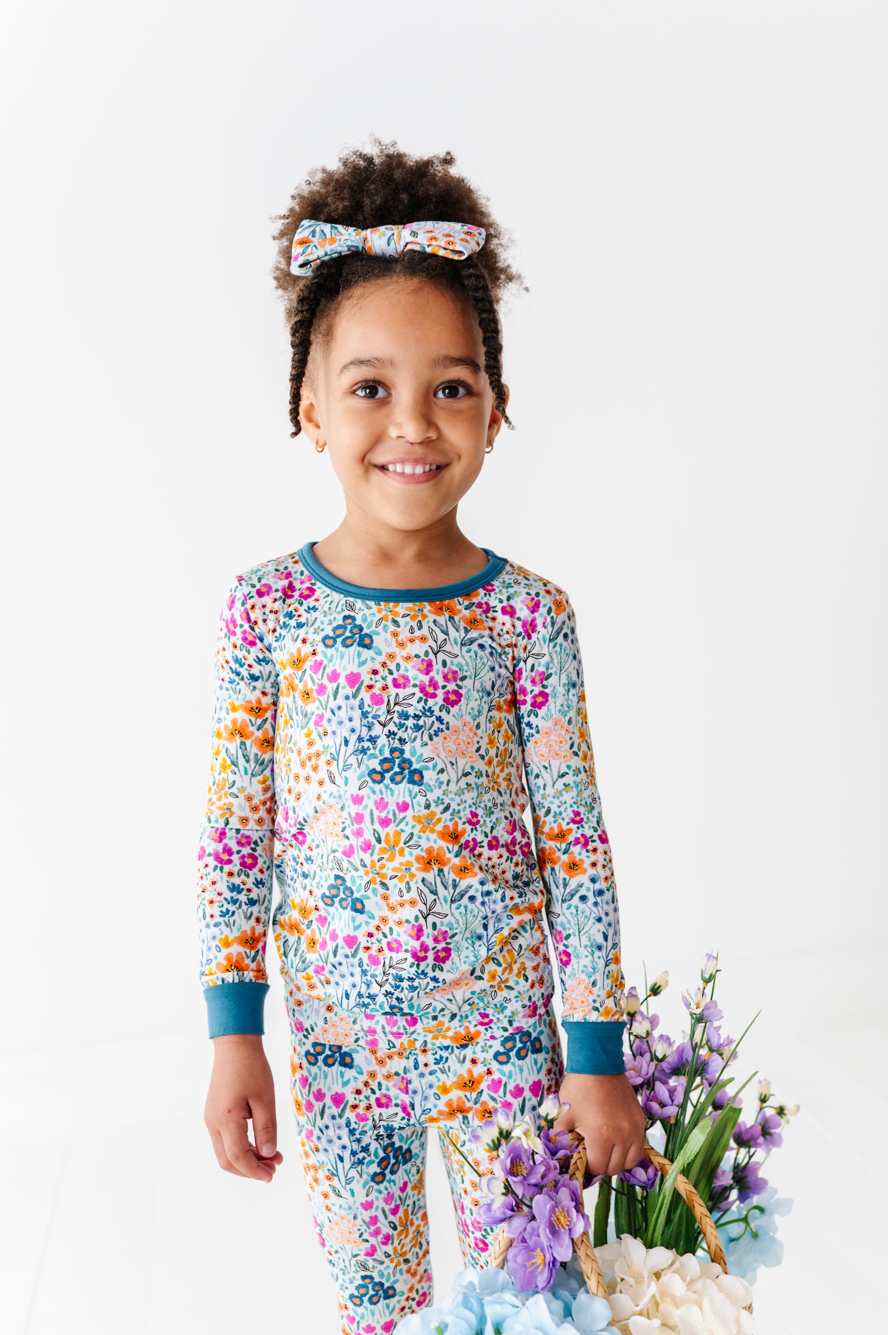 A young child in Jammie Session's Whispering Wildflowers Long Sleeve PJ's with a matching floral headband smiles indoors beside a basket of colorful flowers.