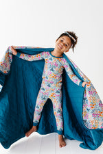 A smiling child in floral pajamas holds open the Jammie Session Whispering Wildflowers Toddler Quilted Blanket (2.5 Tog, machine washable bamboo) against a white background.
