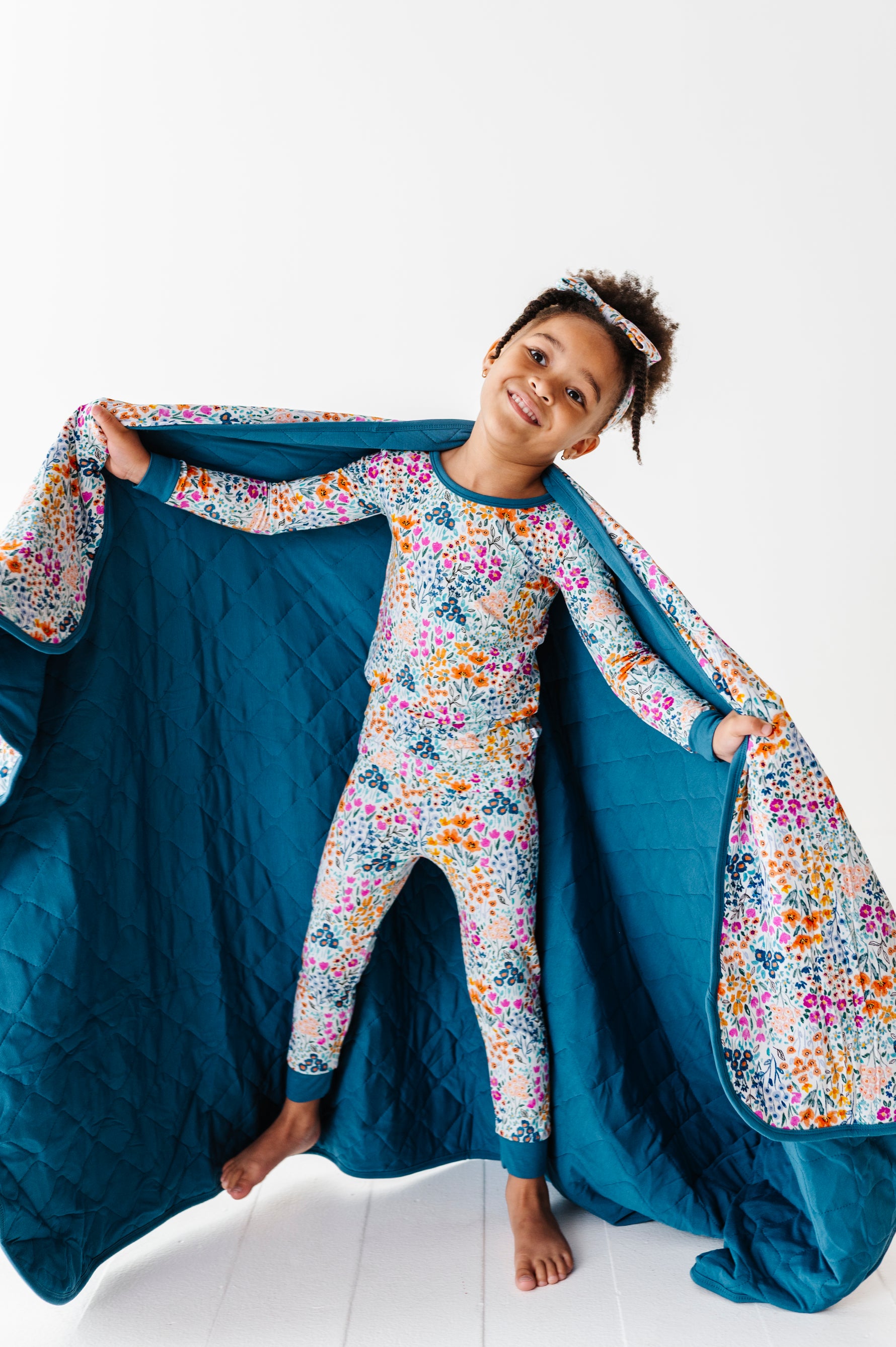 A smiling child in floral pajamas holds open the Jammie Session Whispering Wildflowers Toddler Quilted Blanket (2.5 Tog, machine washable bamboo) against a white background.