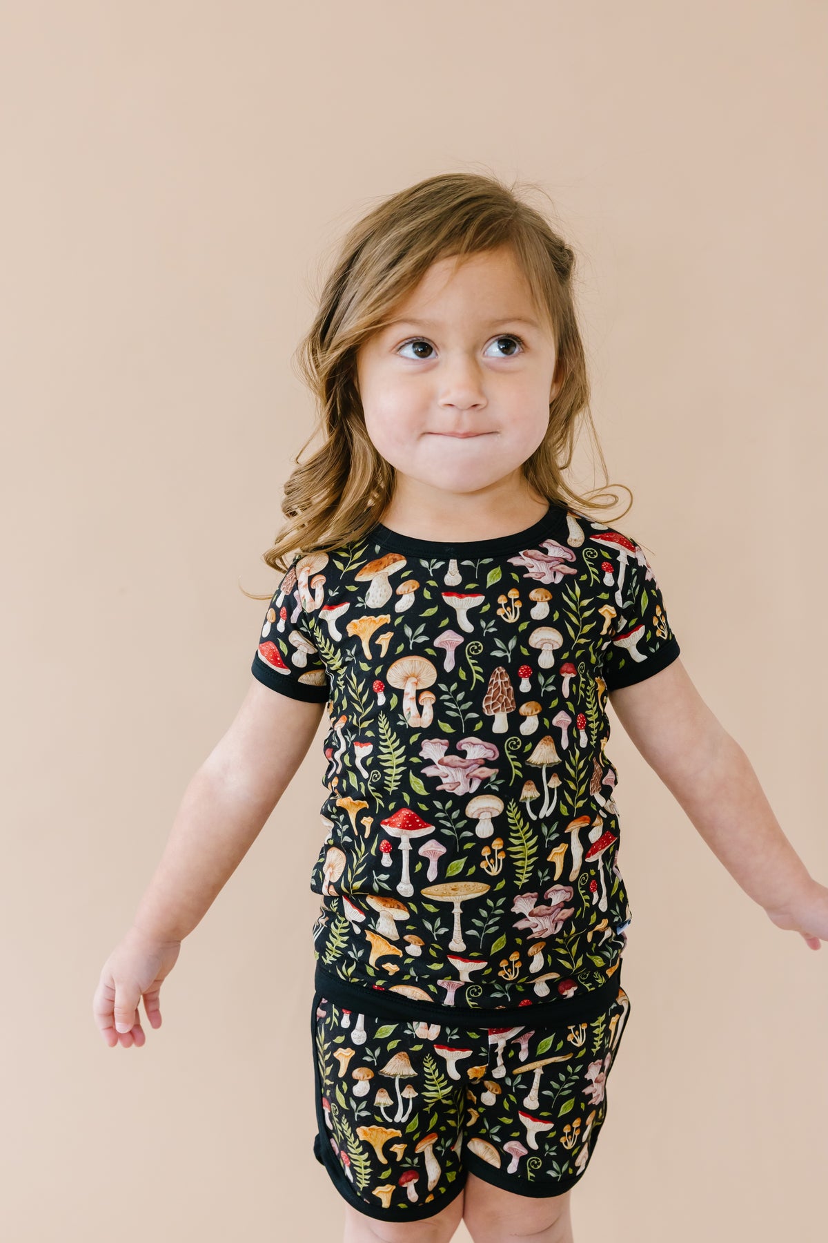 A young child in Jammie Session’s Mystic Mushrooms Short Pajama Set stands against a beige background—these breathable bamboo pajamas feature a colorful mushroom pattern and are gentle on sensitive skin.