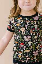 A young child with light brown wavy hair wears the Jammie Session Mystic Mushrooms Short Pajama Set, featuring a black, hypoallergenic kids' shirt with a colorful mushroom and fern pattern, facing forward with a neutral expression.