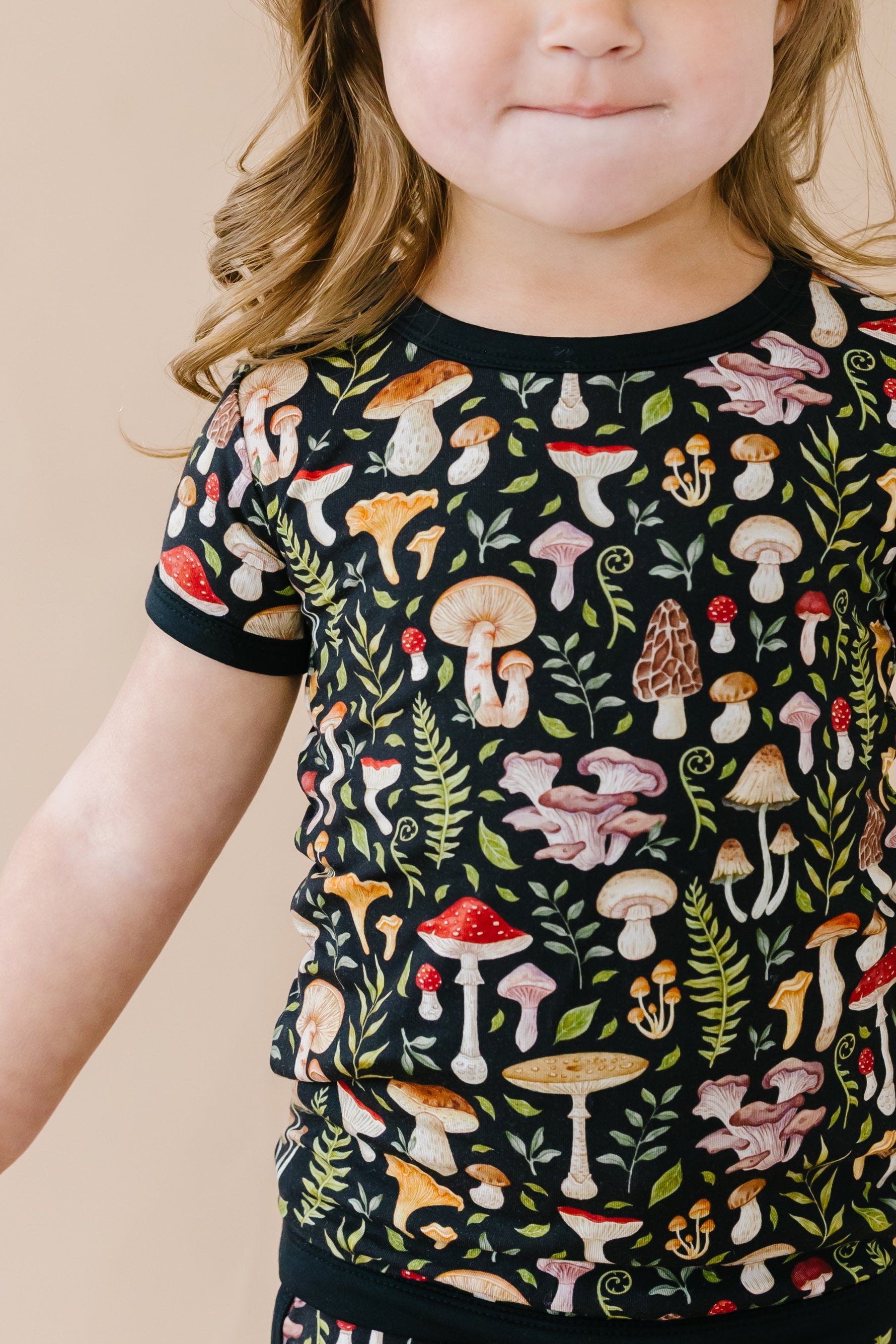 A young child with light brown wavy hair wears the Jammie Session Mystic Mushrooms Short Pajama Set, featuring a black, hypoallergenic kids' shirt with a colorful mushroom and fern pattern, facing forward with a neutral expression.