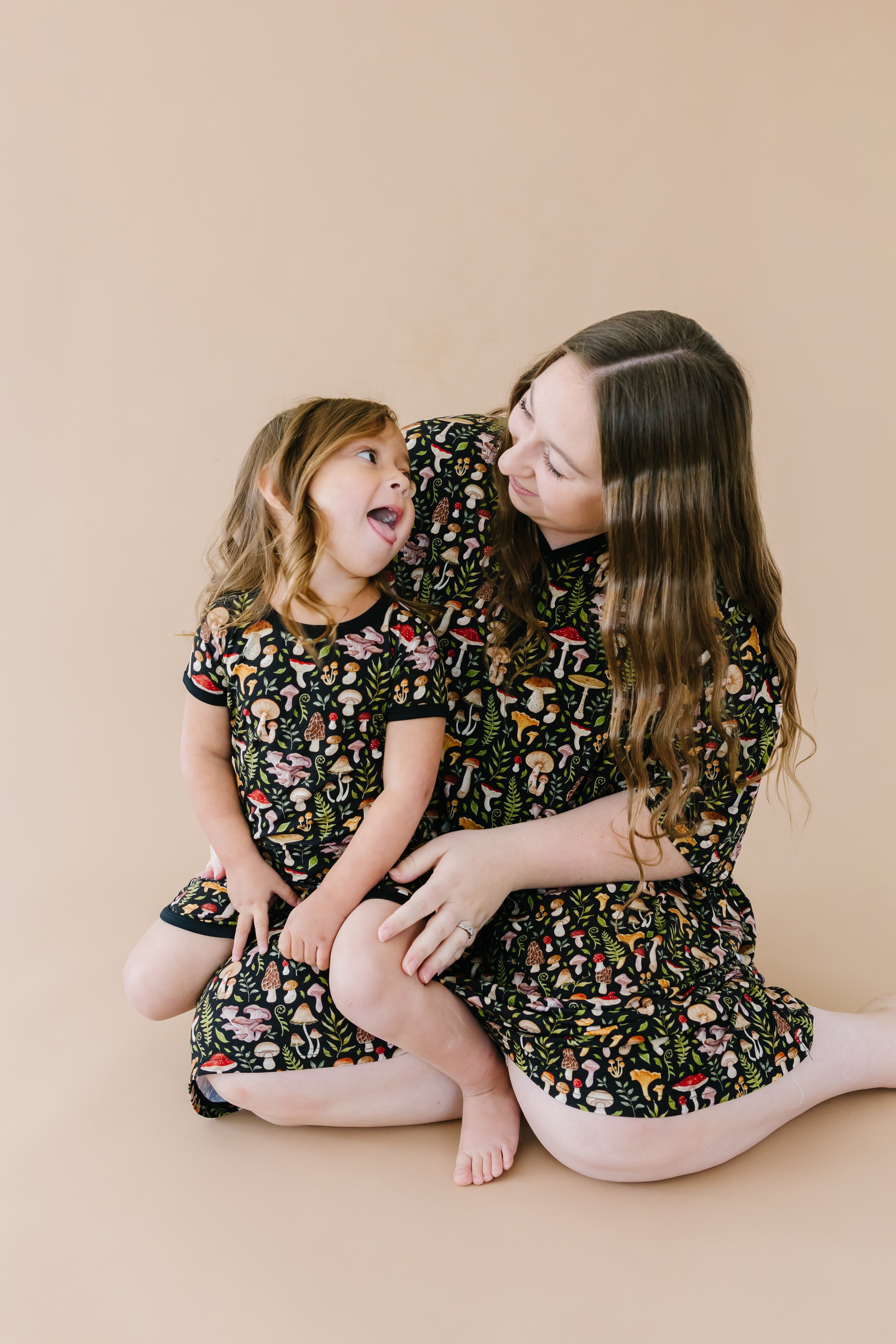 A woman and a young girl sit on the floor against a beige background, smiling at each other in matching Jammie Session Mystic Mushrooms Short Pajama Sets—stylish, hypoallergenic kids' clothing with colorful patterns.