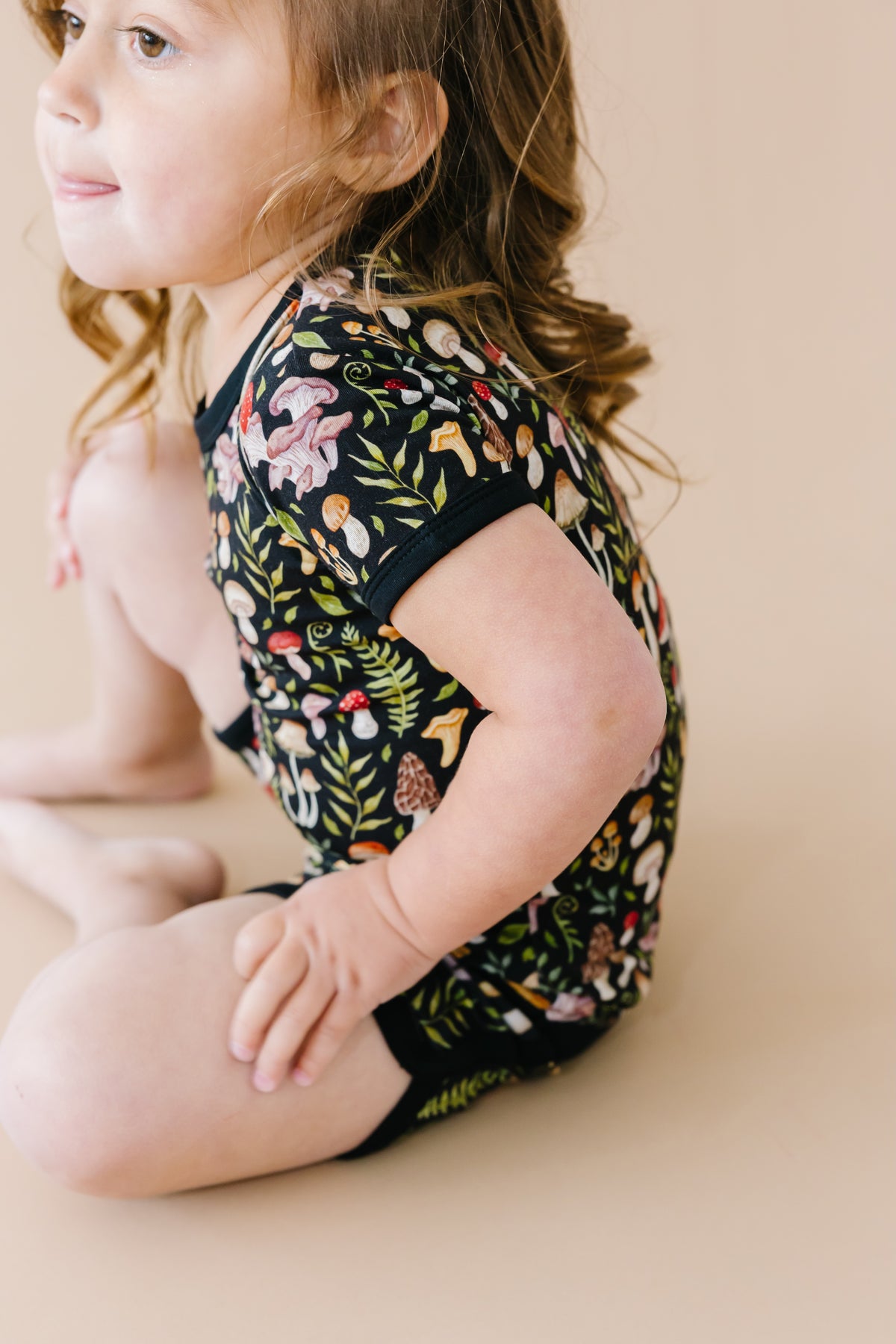A young child with brown hair sits on a beige surface, wearing the Jammie Session Mystic Mushrooms Short Pajama Set—breathable kids’ sleepwear with a vibrant nature-inspired print for ultimate comfort.