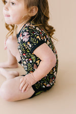 A young child with brown hair sits on a beige surface, wearing the Jammie Session Mystic Mushrooms Short Pajama Set—breathable kids’ sleepwear with a vibrant nature-inspired print for ultimate comfort.