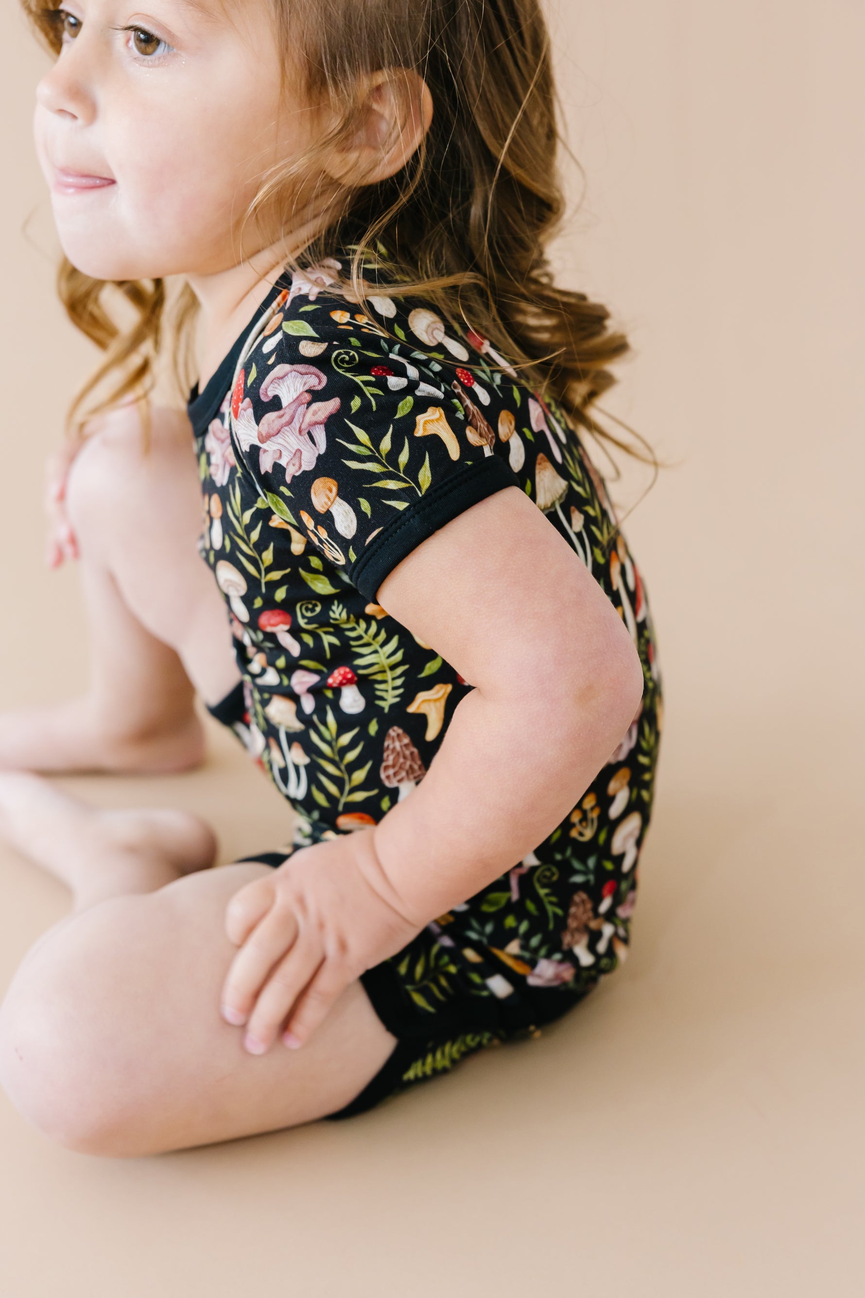 A young child with brown hair sits on a beige surface, wearing the Jammie Session Mystic Mushrooms Short Pajama Set—breathable kids’ sleepwear with a vibrant nature-inspired print for ultimate comfort.