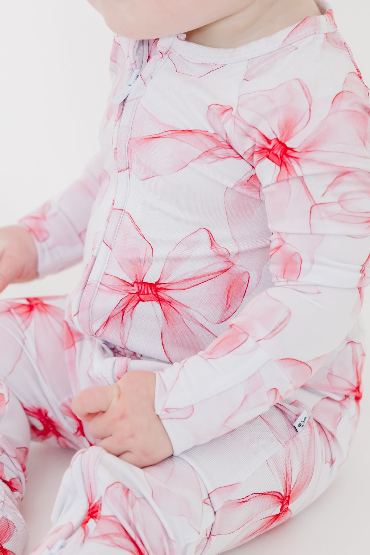 A baby wears the Pretty in Bows Romper by Jammie Session, featuring white long sleeves with bold pink and red floral patterns, seated and facing slightly to the side.