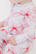 A baby wears the Pretty in Bows Romper by Jammie Session, featuring white long sleeves with bold pink and red floral patterns, seated and facing slightly to the side.