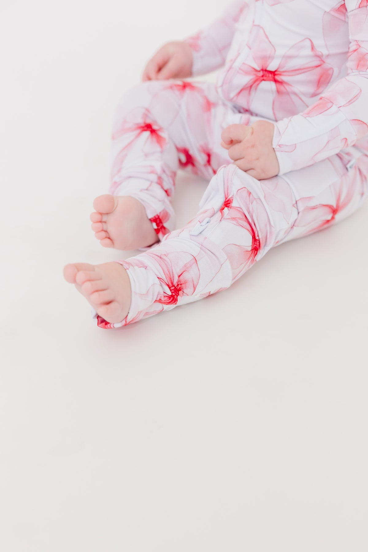 A baby wears the Jammie Session Pretty in Bows Romper, a white long sleeve outfit with pink and red floral patterns, sitting on a white surface with feet extended and hands resting on their legs.
