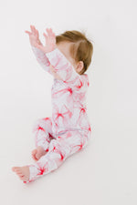 A baby in the Jammie Session Pretty in Bows Romper, featuring a white and pink floral pattern with long sleeves, sits on the floor with arms raised against a white background.
