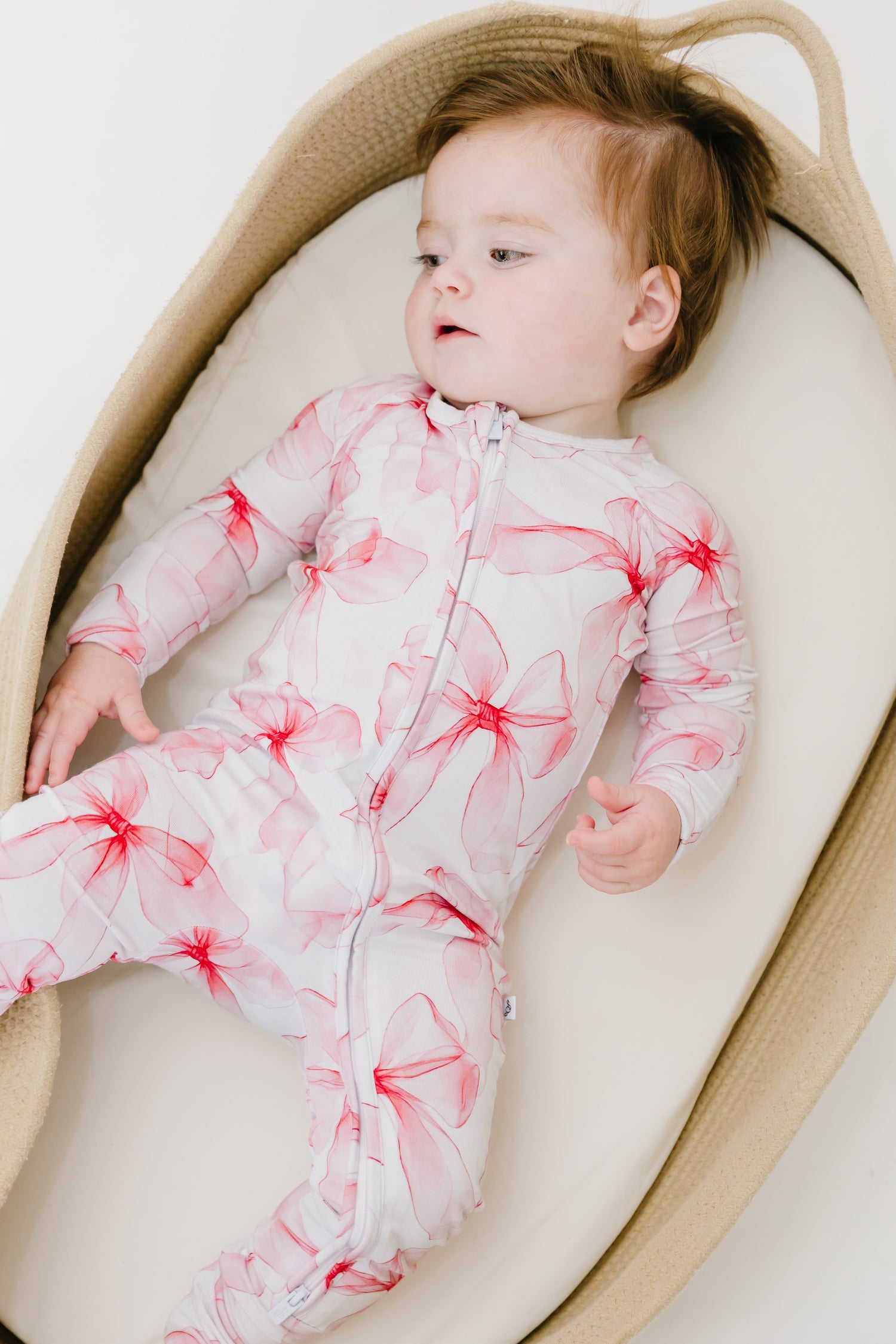 A baby cozies up in a basket, wearing the Jammie Session Pretty in Bows Romper—a white and pink floral-patterned long sleeve outfit—looking off to the side.