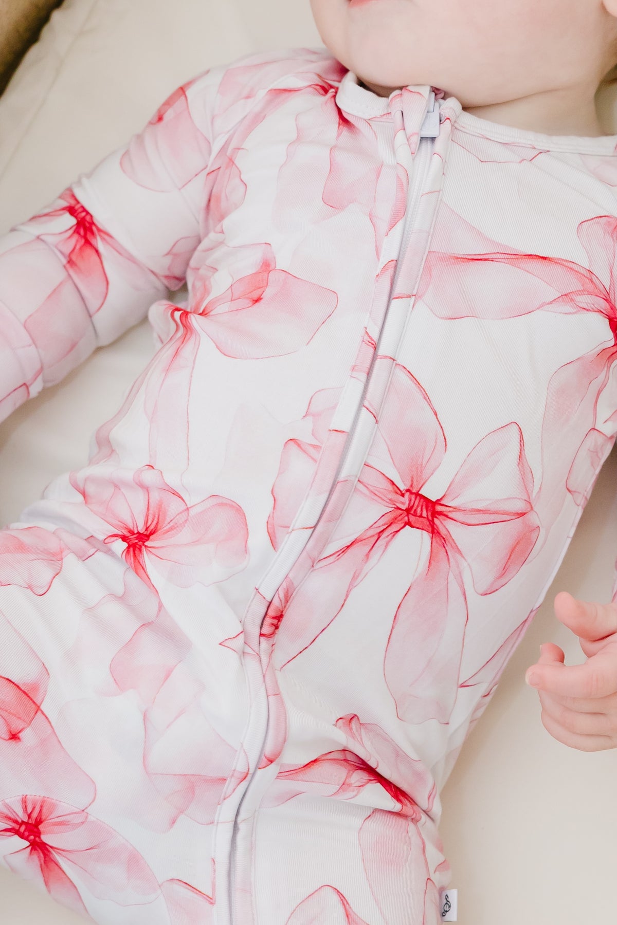 A baby in the Jammie Session Pretty in Bows Romper—a white bamboo outfit with pink floral patterns—lies down with one hand partially visible.