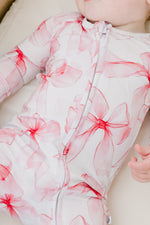 A baby in the Jammie Session Pretty in Bows Romper—a white bamboo outfit with pink floral patterns—lies down with one hand partially visible.