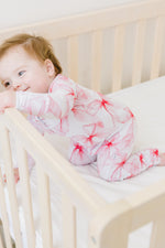 A baby in a white crib wears the Pretty in Bows Romper by Jammie Session, featuring pink floral patterns and long sleeves, holding the crib rail and looking to the side.