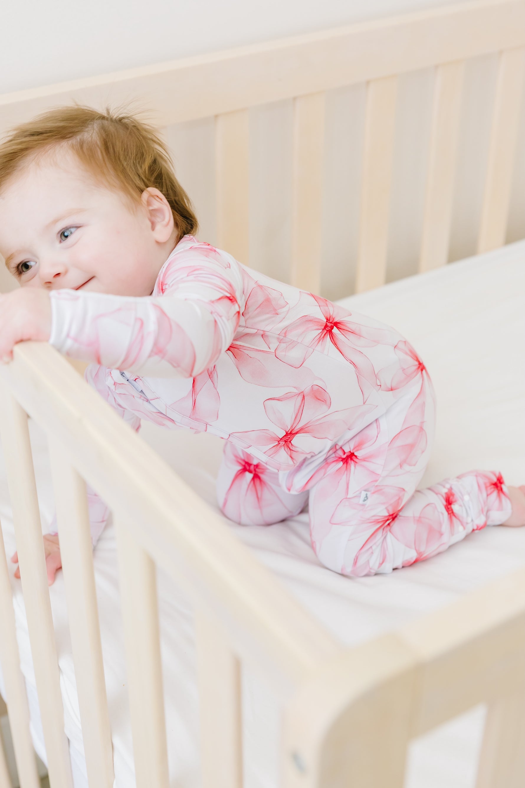 A baby in a white crib wears the Pretty in Bows Romper by Jammie Session, featuring pink floral patterns and long sleeves, holding the crib rail and looking to the side.