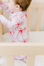 A baby in the Jammie Session Pretty in Bows Romper stands inside a light wooden crib, holding the rail.
