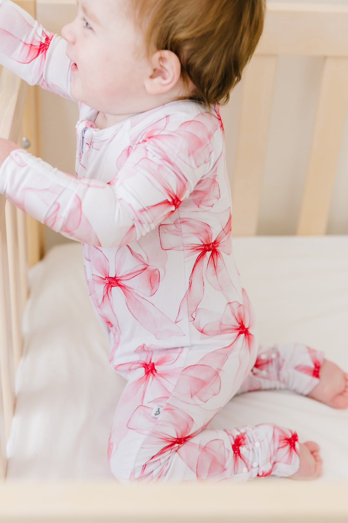 A baby kneels in a white crib, holding the railings while wearing the Jammie Session Pretty in Bows Romper, a long sleeve outfit adorned with pink and red flower patterns.