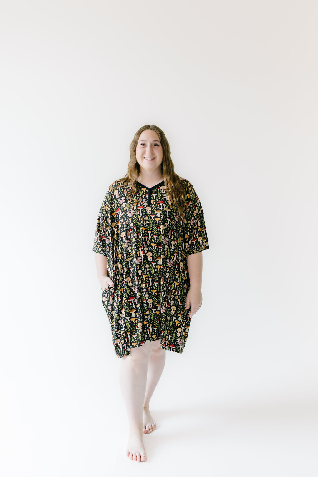 A woman with long hair stands barefoot on a white background, smiling at the camera in a Jammie Session Mystic Mushrooms Women's Muumuu Lounge Tee, a loose, patterned dress with short sleeves and pockets (ONE SIZE FITS XS - XXL).