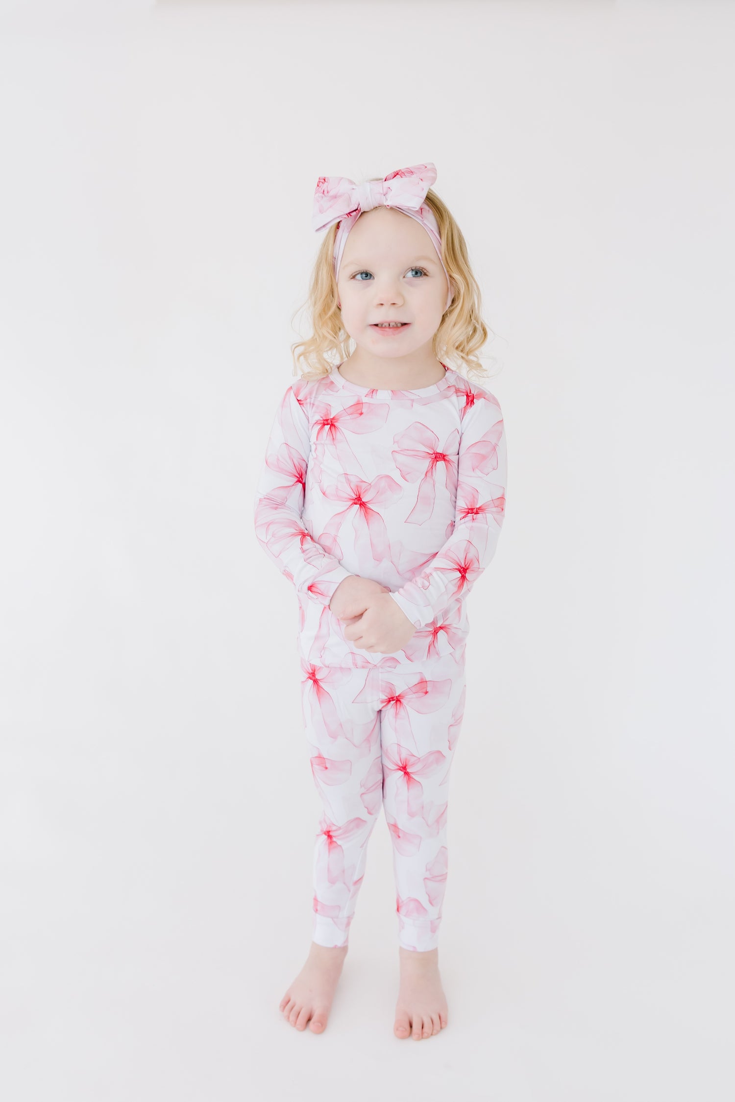 A young child stands barefoot against a white background, wearing Jammie Session's Pretty in Bows Long Sleeve PJ's—floral bamboo pajamas with a matching bow headband, gentle on skin and perfect for sensitive sleepers.