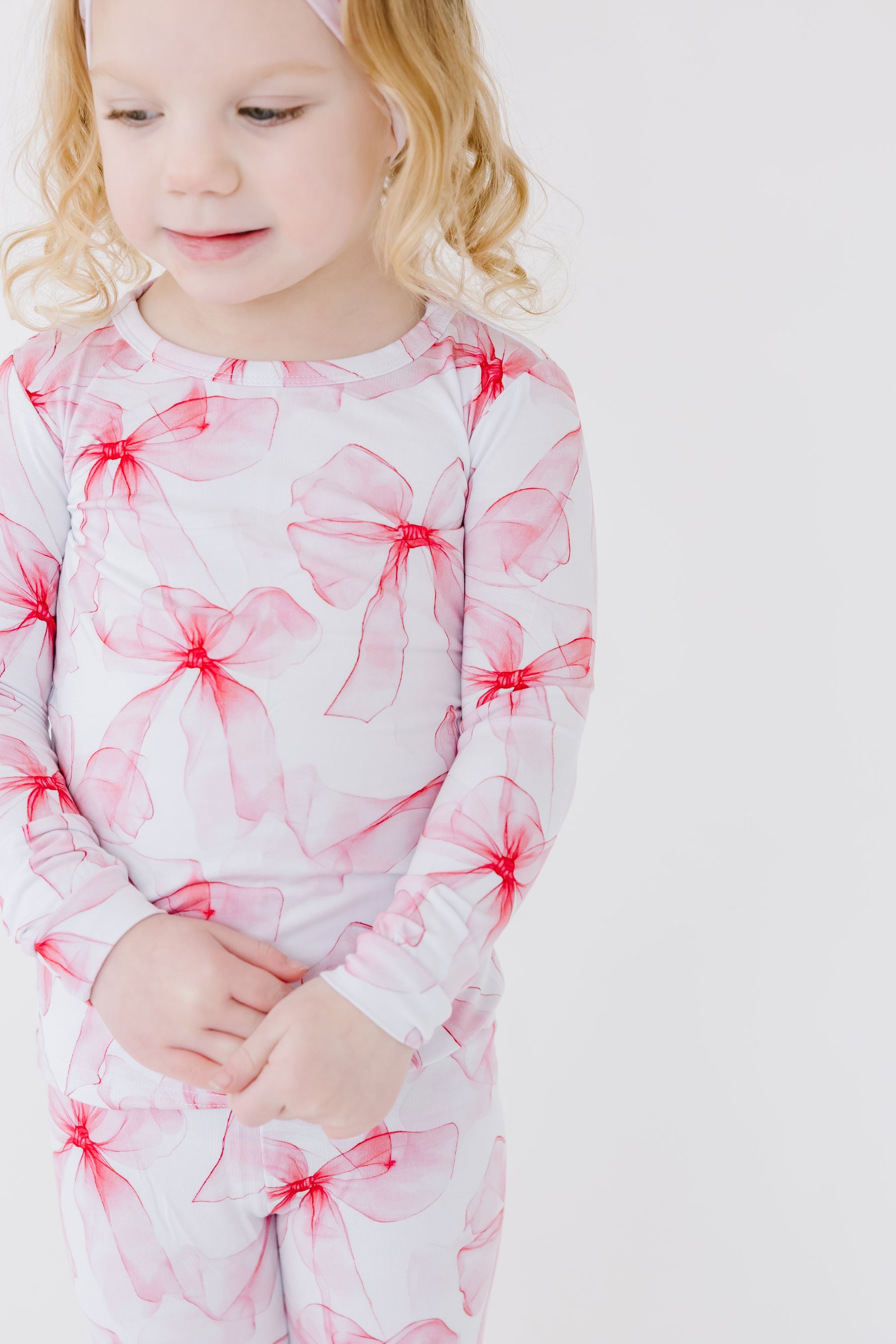 A young blonde child wears Jammie Session’s Pretty in Bows Long Sleeve PJ's, featuring red bow patterns, standing against a plain white background.