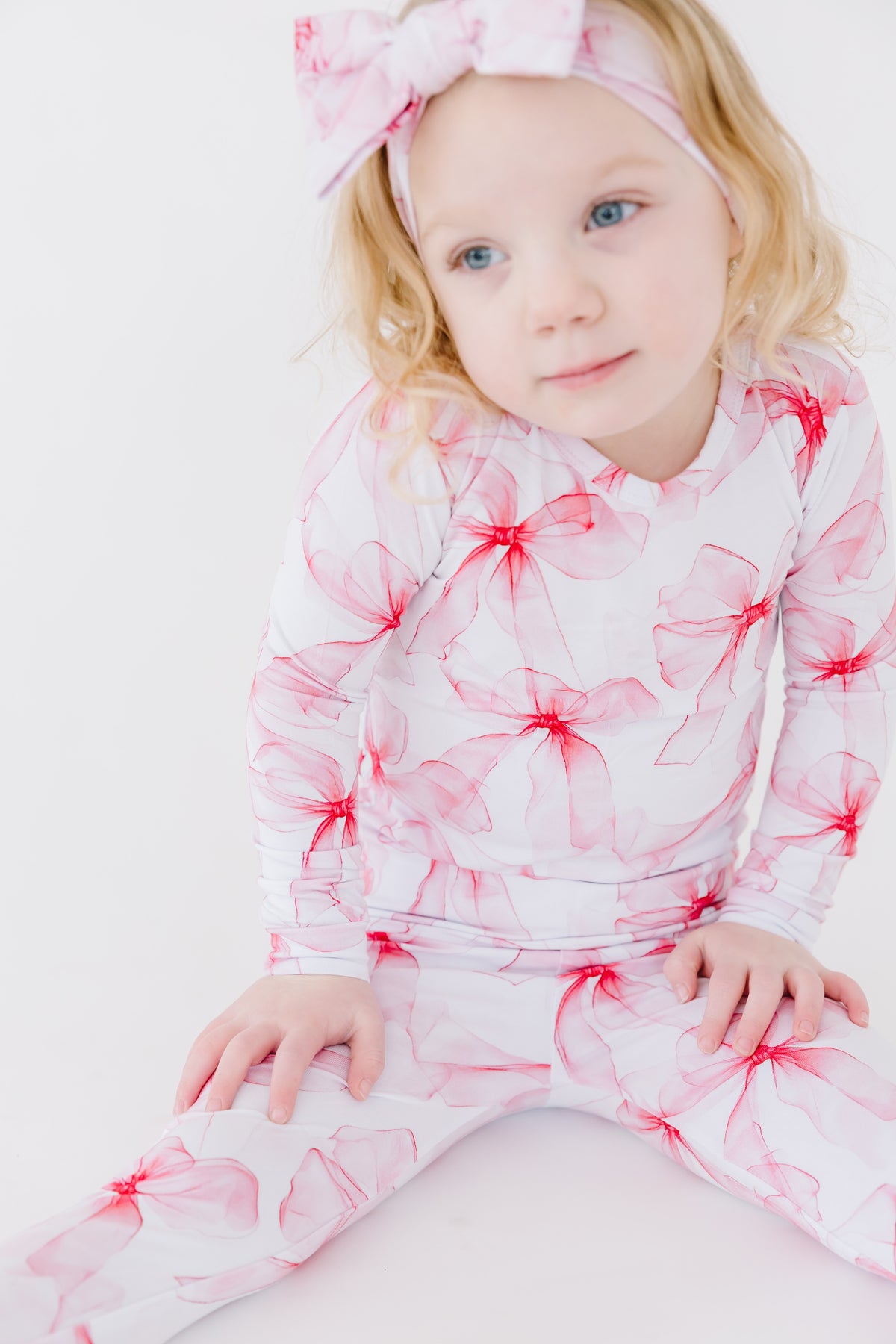 A young child in Jammie Session's Pretty in Bows Long Sleeve PJ's—a white and pink floral set with a matching headband—sits on the floor against a plain white background.