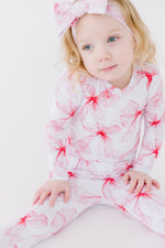 A young child in Jammie Session's Pretty in Bows Long Sleeve PJ's—a white and pink floral set with a matching headband—sits on the floor against a plain white background.