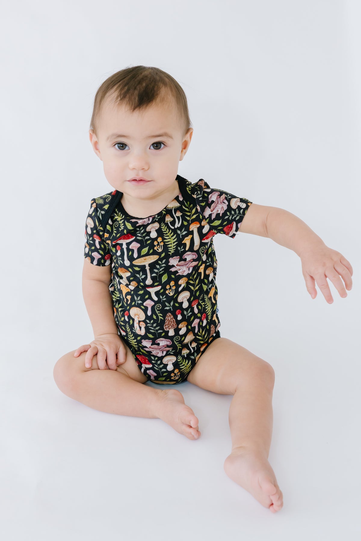 A baby wearing the Jammie Session Mystic Mushrooms Short Sleeve Bodysuit, featuring a colorful mushroom pattern, sits on a white background with one arm extended.